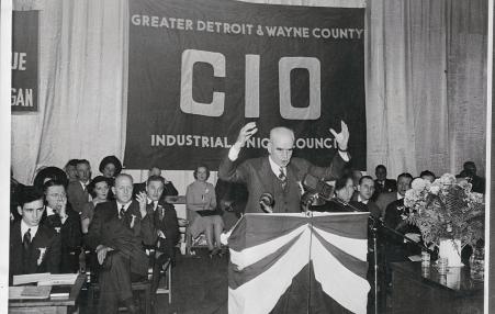 A speaker at a podium with a full house of workers behind him and a huge banner CIO